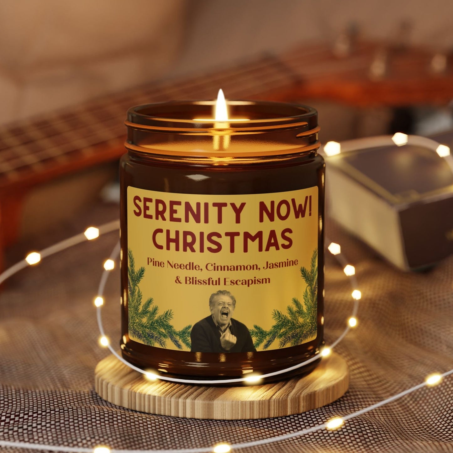 Candle in a jar labeled 'Serenity Now Christmas' with decorative lights and guitar in the background 9oz